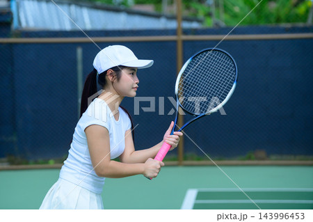 Young Asian Woman Playing Tennis on Outdoor Court in White Sportswear Holding Racket Ready Position Young Asian Woman Playing Tennis on Outdoor Court in White Sportswear Holding Racket Ready Position 143996453