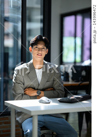 Young Asian Businessman Sitting at Office Desk with Arms Crossed, Confident Smile, Gray Blazer Young Asian Businessman Sitting at Office Desk with Arms Crossed, Confident Smile, Gray Blazer 143996472