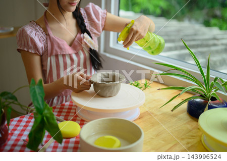 Cropped shot Woman Shaping Clay Bowl on Pottery Wheel While Spraying Water in Bright Studio 143996524