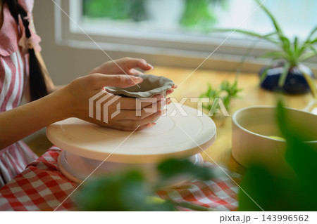 Young Woman Shaping Clay by Hand on Pottery Turntable at Home Studio with Green Plants 143996562