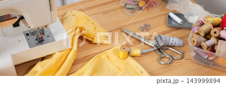 Yellow trousers lie on a wooden work table as their hem is shortened with scissors, a measuring tape, pins, and sewing tools during a clothing alteration, banner 143999894