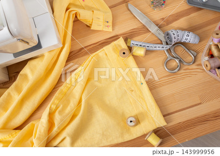 Yellow trousers lie on a wooden work table as their hem is shortened with scissors, a measuring tape, pins, and sewing tools during a clothing alteration Yellow trousers lie on a wooden work table as their hem is shortened with scissors, a measuring tape, pins, and sewing tools during a clothing alteration 143999956