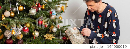 A 53-year-old man kneels by a Christmas tree in his home on Christmas Day. He examines a gift carefully as holiday decorations surround him. The moment is filled with family joy and cheer 144000019