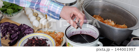 Hands add dried hibiscus, also called Sudanese rose, to a pot of dye for Easter eggs in a home kitchen, with turmeric used as another natural dye, banner 144001997