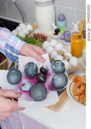 Hands dye eggs with red cabbage in pots on a kitchen stove, then place the eggs on a plate during Easter preparations at home 144002002