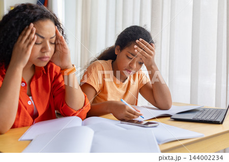 Stressed black mother and tired little daughter holding heads while doing homework Stressed black mother and tired little daughter holding heads while doing homework 144002234