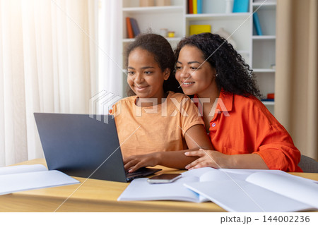 Smiling black mother and daughter sitting together at desk using laptop at home 144002236
