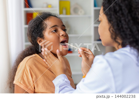 Black female doctor examining young girls throat using wooden tongue depressor 144002238