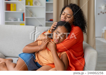 Black mother comforting her upset daughter while sitting on sofa at home 144002256