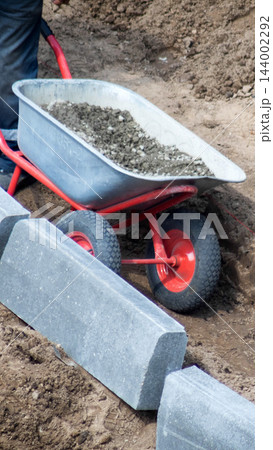 Construction worker prepares ground for road curbing installation, pushing wheelbarrow full of gravel near concrete blocks, person leg visible performing work, outdoors day vertical photo 144002292