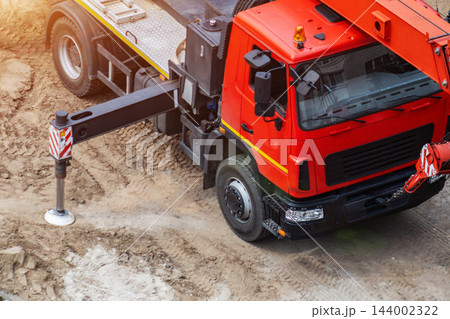 Red truck crane with extended outrigger stabilizer on dusty construction ground, heavy vehicle for lifting material on building site, engineering machinery work, copy space 144002322