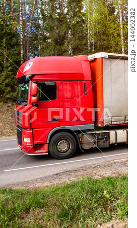 Red semi-truck with white cargo trailer moves quickly on asphalt road next to green forest under sunny sky, blurring background for sense of speed, vertical photo Red semi-truck with white cargo trailer moves quickly on asphalt road next to green forest under sunny sky, blurring background for sense of speed, vertical photo 144002382