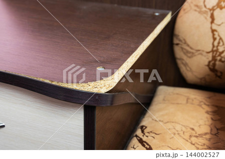 Close up view of damaged particle board shelf edge with brown veneer and exposed compressed wood fibers showing wear and tear of furniture material 144002527