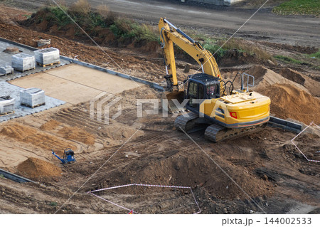 Yellow tracked excavator works at construction site during tile laying process with sand mounds pallets of paving stones ground preparation and compacting equipment 144002533