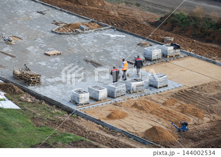 High angle view of construction workers installing concrete paving stones on pedestrian sidewalk with pallets of tiles, sand base, and compacting equipment outdoors. 144002534