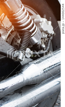 Close-up detail of wet chrome exhaust pipes and rear suspension of motorcycle, highlighting shock absorber, brake disc, and wheel spokes with dynamic sun flare on metal surface. 144002544