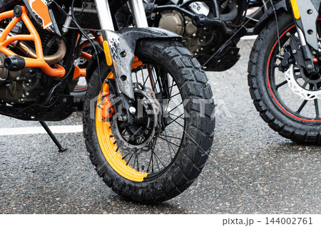 Close up view front wheel orange black adventure motorcycle parked wet asphalt street, textured off-road tire visible brake disc caliper components, bright day light Close up view front wheel orange black adventure motorcycle parked wet asphalt street, textured off-road tire visible brake disc caliper components, bright day light 144002761