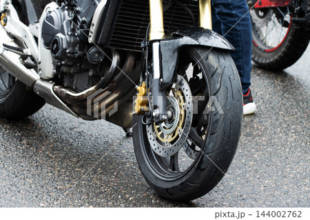 Close up view of front wheel motorcycle, tire tread on wet asphalt, golden brake caliper, front disc brake system, engine detail, exhaust pipes, shiny suspension forks, showcasing modern vehicle 144002762
