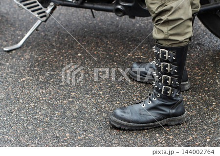 Close up of person legs wearing heavy black leather spiked boots and olive green pants, standing beside parked motorcycle footpeg on wet asphalt road surface, gritty biker fashion 144002764