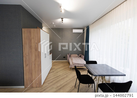 Modern studio apartment interior showing compact living room space with contemporary dining table, black chairs, wardrobe cabinet, sofa bed, parquet floor, tulle curtains Modern studio apartment interior showing compact living room space with contemporary dining table, black chairs, wardrobe cabinet, sofa bed, parquet floor, tulle curtains 144002791