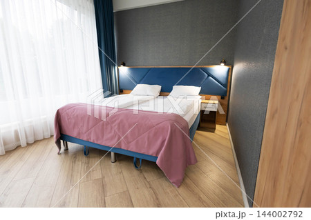modern hotel bedroom interior featuring comfortable double bed with blue headboard, stylish pink throw blanket, white pillows, nightstand, wooden floor, and large window 144002792