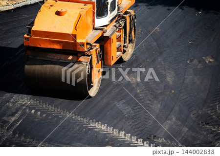 Orange asphalt paving roller compacts fresh black asphalt during road construction activity, creating smooth surface for new infrastructure development work under bright sunlight. Copy space 144002838