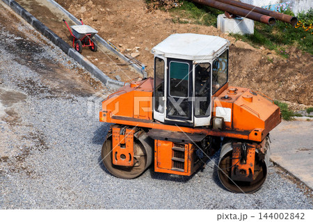 Aerial view of large orange and white asphalt paving roller compacting layer of crushed stone at new road construction site. Equipment stands on uneven gravel surface with concrete curb and 144002842