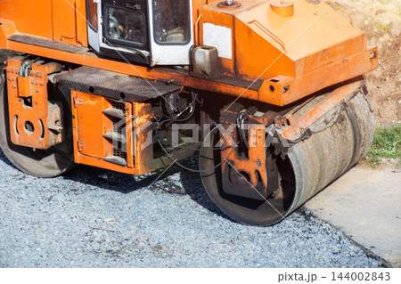 Orange road roller working construction site, compacting gravel new roadbed. Heavy machinery used civil engineering project infrastructure development. 144002843