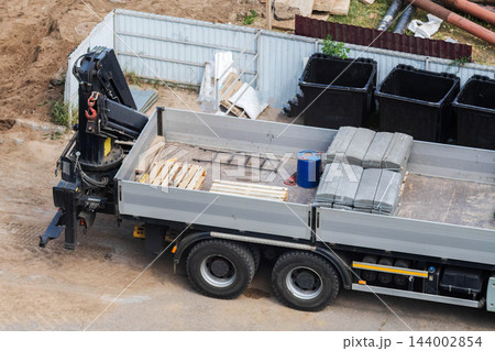 Industrial truck with powerful hydraulic manipulator crane arm positioned at rear, actively unloading heavy concrete road parts and construction materials on a dusty outdoor job site with a fence Industrial truck with powerful hydraulic manipulator crane arm positioned at rear, actively unloading heavy concrete road parts and construction materials on a dusty outdoor job site with a fence 144002854