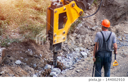 Powerful hydraulic hammer attachment breaking rocky ground at construction site, worker observing progress of excavation activity, heavy equipment operation under bright sunlight. Copy space Powerful hydraulic hammer attachment breaking rocky ground at construction site, worker observing progress of excavation activity, heavy equipment operation under bright sunlight. Copy space 144002910