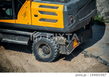 Yellow and dark grey construction machinery, excavator side view with rugged tire on sandy ground, showing rear section of powerful digging equipment for heavy duty work, copy space 144002987
