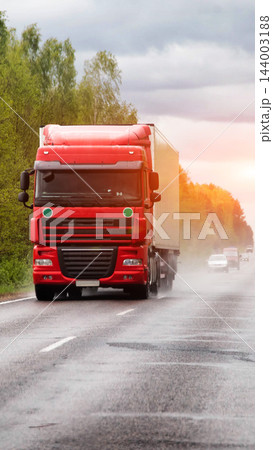 Red semi-truck with white cargo trailer driving on wet asphalt road, water spray from wheels, motion blur effect, cloudy sky with warm sunset light glow over trees, highway transport vertical photo 144003188