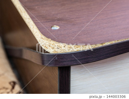Close-up detail of damaged particle board from a wooden cabinet, showing broken edge and peeling surface finish, with a screw securing board, highlighting deterioration of furniture material. Close-up detail of damaged particle board from a wooden cabinet, showing broken edge and peeling surface finish, with a screw securing board, highlighting deterioration of furniture material. 144003306