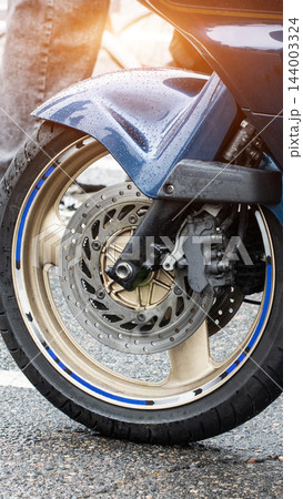 Close-up detail of front wheel of motorcycle on wet asphalt showing disc brake system, blue fender with water droplets, and golden rim with blue arrow pattern side 144003324