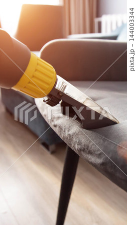 Professional cleaning service worker uses specialized industrial vacuum cleaner machine with yellow nozzle to deep clean upholstered grey fabric sofa furniture for stain removal indoor home vertical 144003344