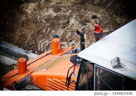 Overhead view of large orange asphalt paving machine, featuring detail of cabin, red warning light, exhaust pipe, and various metal components on construction site dirt ground Overhead view of large orange asphalt paving machine, featuring detail of cabin, red warning light, exhaust pipe, and various metal components on construction site dirt ground 144003446