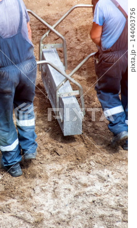 Two construction workers in blue overalls installing road curb using specialized tool, collaborating on a construction site, close up view of hands and equipment, vertical photo 144003756