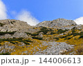 rocky scree path cuts through golden grasses and low shrubs toward pale limestone ridges, with scattered clouds crossing the vivid blue sky 144006613