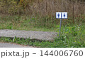 On a metal post amidst the grass, blue-and-white pictograms for men and women restrooms are displayed; the post is situated near a concrete ramp, with roadside thickets in the background. 144006760