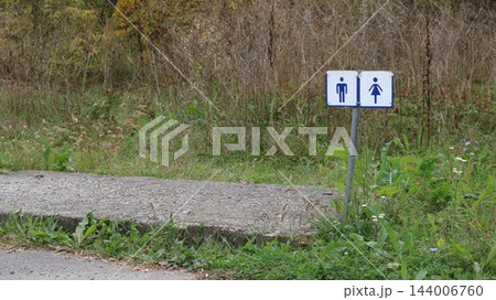 On a metal post amidst the grass, blue-and-white pictograms for men and women restrooms are displayed; the post is situated near a concrete ramp, with roadside thickets in the background. 144006760