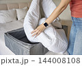 Woman placing white duvet into gray fabric storage box on bed 144008108