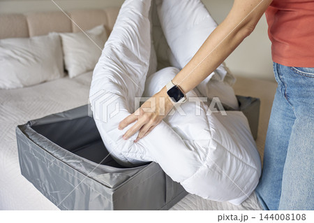 Woman placing white duvet into gray fabric storage box on bed Woman placing white duvet into gray fabric storage box on bed 144008108