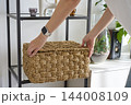 Close up of woman placing large woven wicker storage basket with lid on shelf 144008109