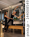 A Pilates instructor supports a client through a side stretch on a trapeze table in a modern studio 144008549
