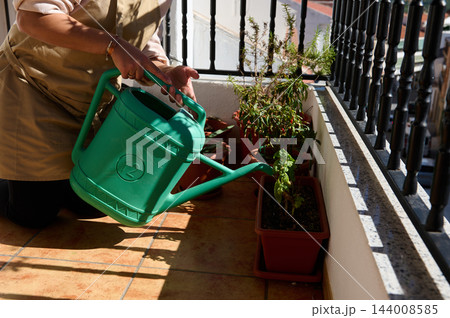 Woman waters potted plants with a green watering can on a sunny tiled apartment balcony, tending a terracotta planter among containers. Her gardening routine supports plant care, urban living, and 144008585