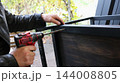 The hands of a craftsman drilling a hole into a dark wooden board attached to a black metal frame. The process of assembling loft-style furniture takes place outdoors. 144008805