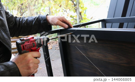 The hands of a craftsman drilling a hole into a dark wooden board attached to a black metal frame. The process of assembling loft-style furniture takes place outdoors. 144008805