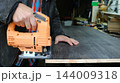 A man hands carefully guide an orange electric jigsaw along a smooth wooden board. The tool makes a clean cut through the solid wood, which is secured to a workbench in a workshop. 144009318