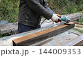 A man wearing a leather jacket and work gloves is working on metal outdoors. Using a blue angle grinder, he cleans and sands the surface of a long, rectangular-profile metal pipe. 144009353