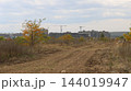 In the background, multi-story concrete residential buildings are under construction, with two tower cranes rising above them. In the foreground lies a spacious, empty plot of land with a dirt road. 144019947